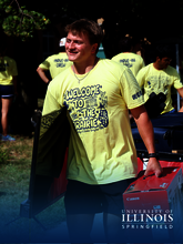 Young man carrying a box, wearing a yellow t-shirt, smiling outdoors.
