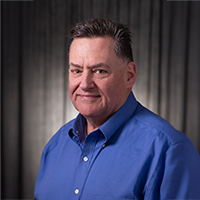 Matthew Scrivano, a middle-aged man in a blue shirt against a dark background.