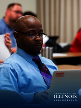 Man in a blue shirt and glasses works on a laptop in a classroom setting.