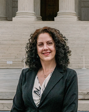 Woman in a suit smiling in front of a building with large steps.