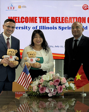 Three people smiling, holding teddy bears, with flags and flowers on a table.