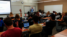 Classroom with a teacher lecturing and students seated, some raising hands.