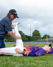 Baseball coach helps player stretch leg on field, cloudy sky.