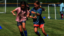 Two girls running on a soccer field, smiling and competing closely.