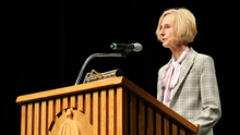 Blonde woman speaking at a wooden podium with a microphone.