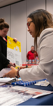 Two people interacting at a busy indoor booth, exchanging documents.