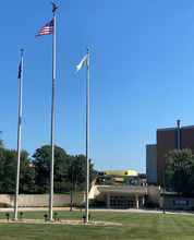 Three tall flagpoles with flags, clear blue sky, and a building in the background.