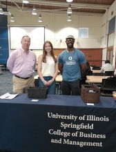 Three people standing behind a university information table indoors.