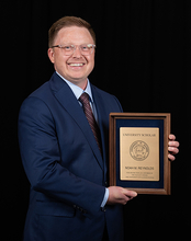 Man in suit holding a wooden plaque, smiling against a black background.