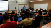 Students in a classroom, some raising hands as a teacher lectures from the front.