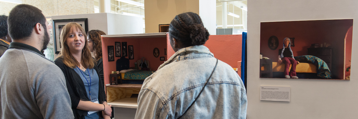 Three people stand talking, looking at a diorama and image of the diorama at a student research symposium.