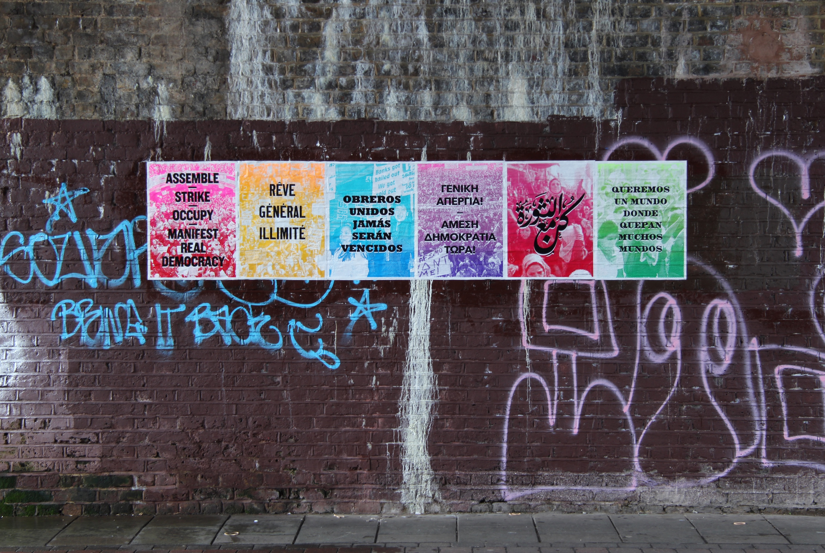 Photo depicts 6 brightly colored screen prints with black text in various languages. All in protest of war. All displayed on a brick wall covered in graffiti.