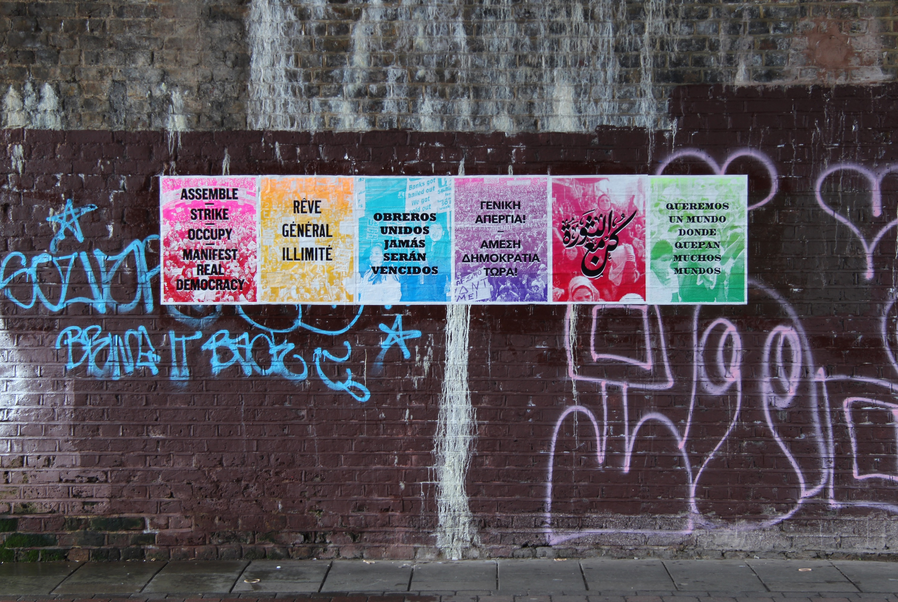 Colorful posters on a graffiti-covered brick wall in a dimly lit alley.