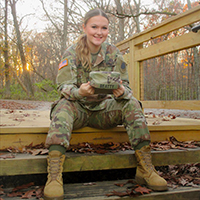 A smiling soldier sits on wooden steps in a forest at sunset holding a cap.