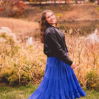 Woman in a bright blue skirt smiles in an autumn field near a pond.