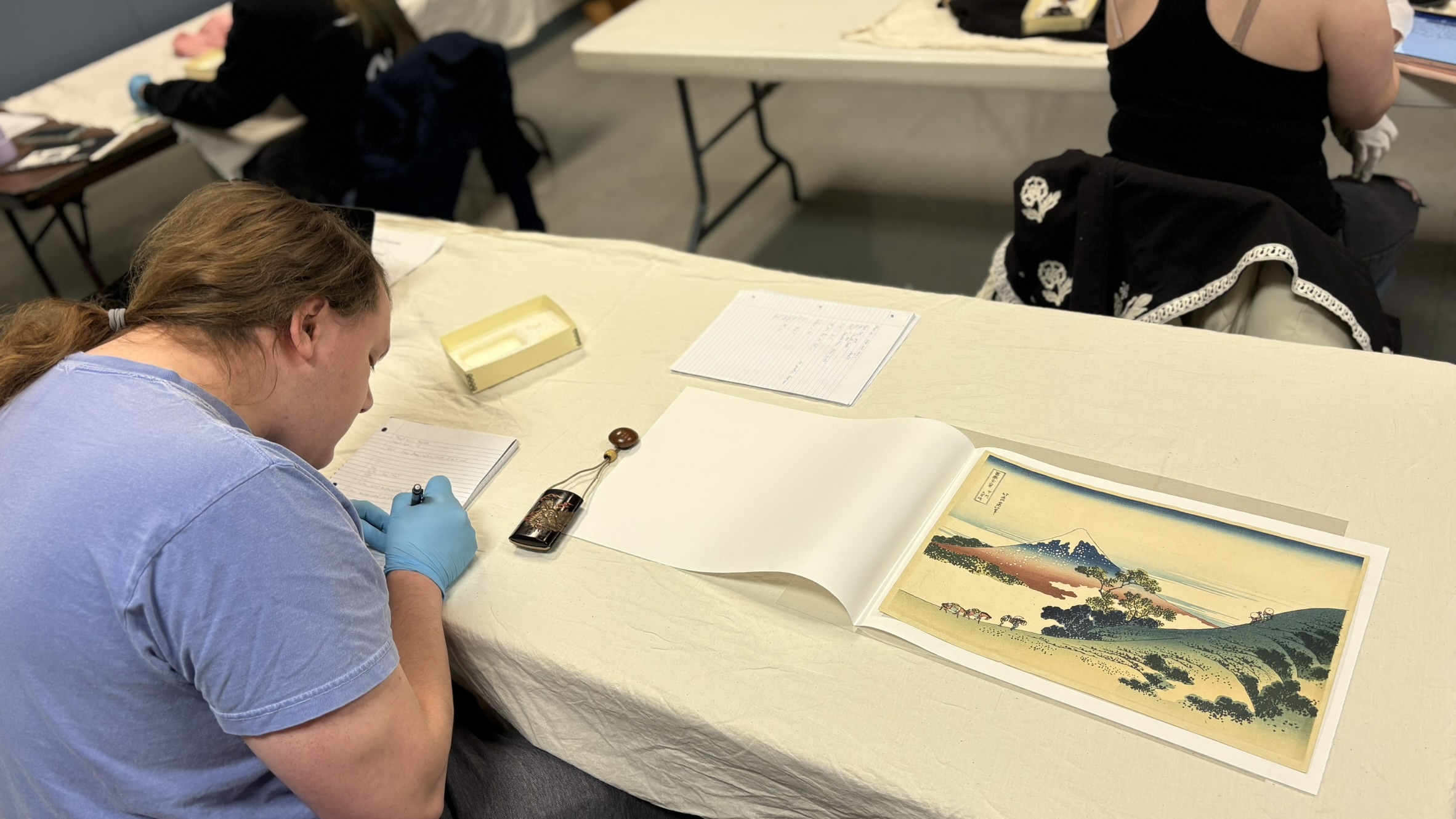 Person in a blue shirt and gloves examines a colorful landscape print at a table, surrounded by notes.