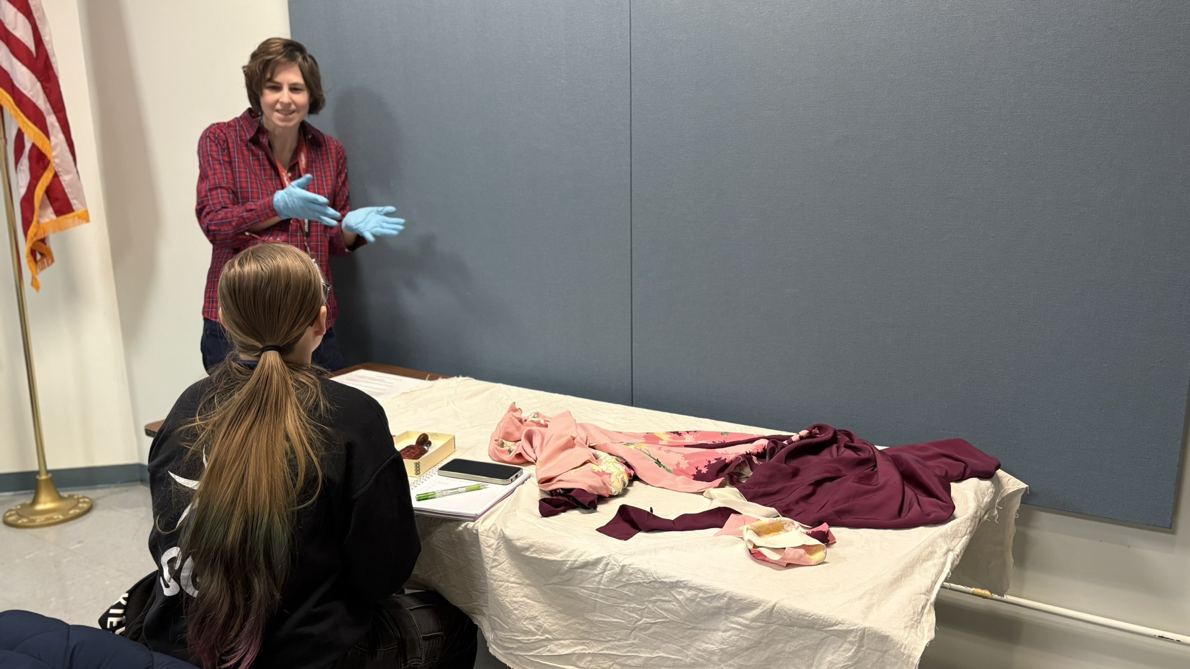 A woman with gloves talks to a seated person, displaying dyed fabrics on a table.