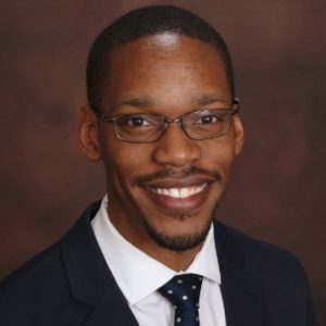 Smiling person in a suit and glasses against dark background.
