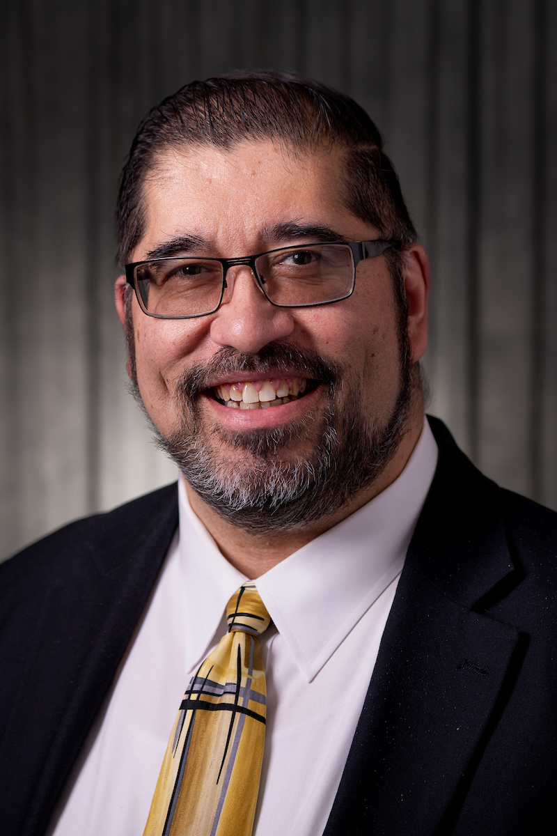 Mark Morten smiles in suit and tie with dark background.