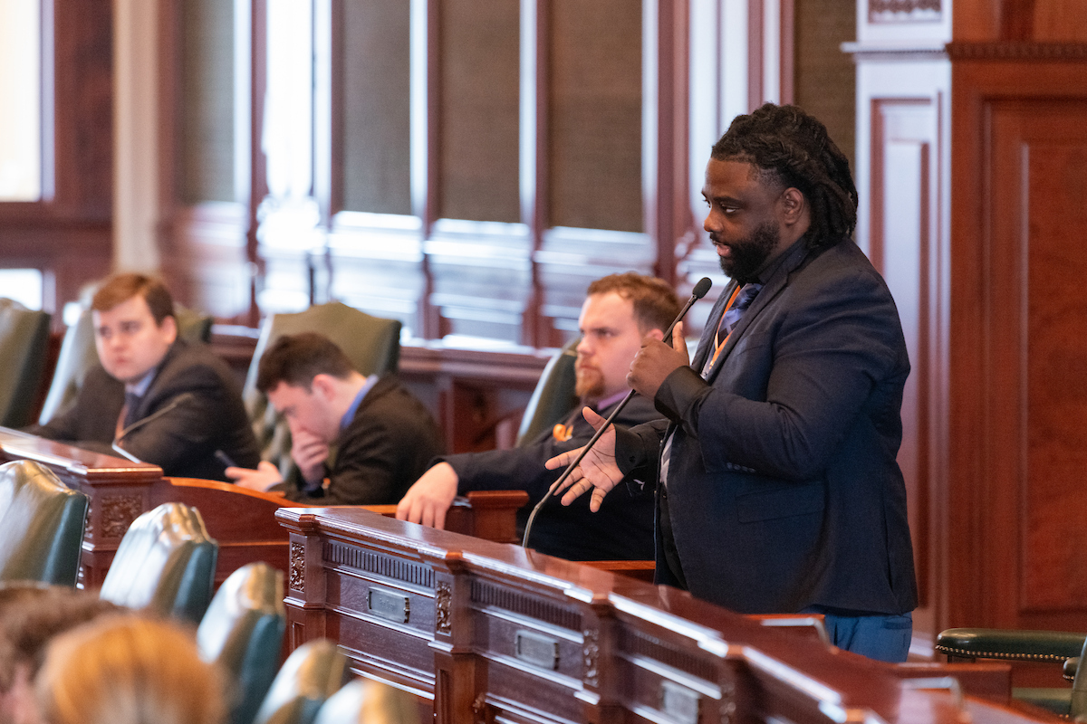 Three students sit in a row in chambers at the Illinois State Capitol while a fourth student stands and speaks into the mic.