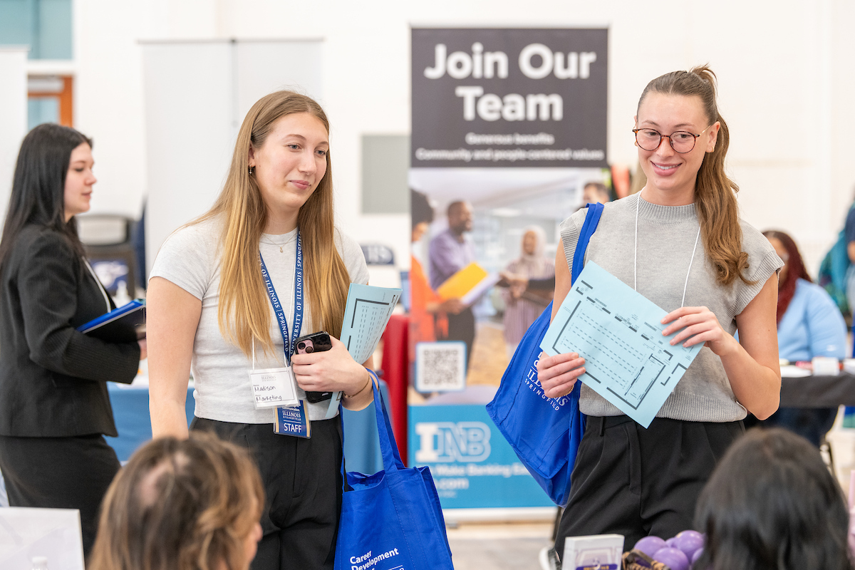Two female students talk with people sitting at a booth at the Career Expo, and a sign behind them says, "Join Our Team."