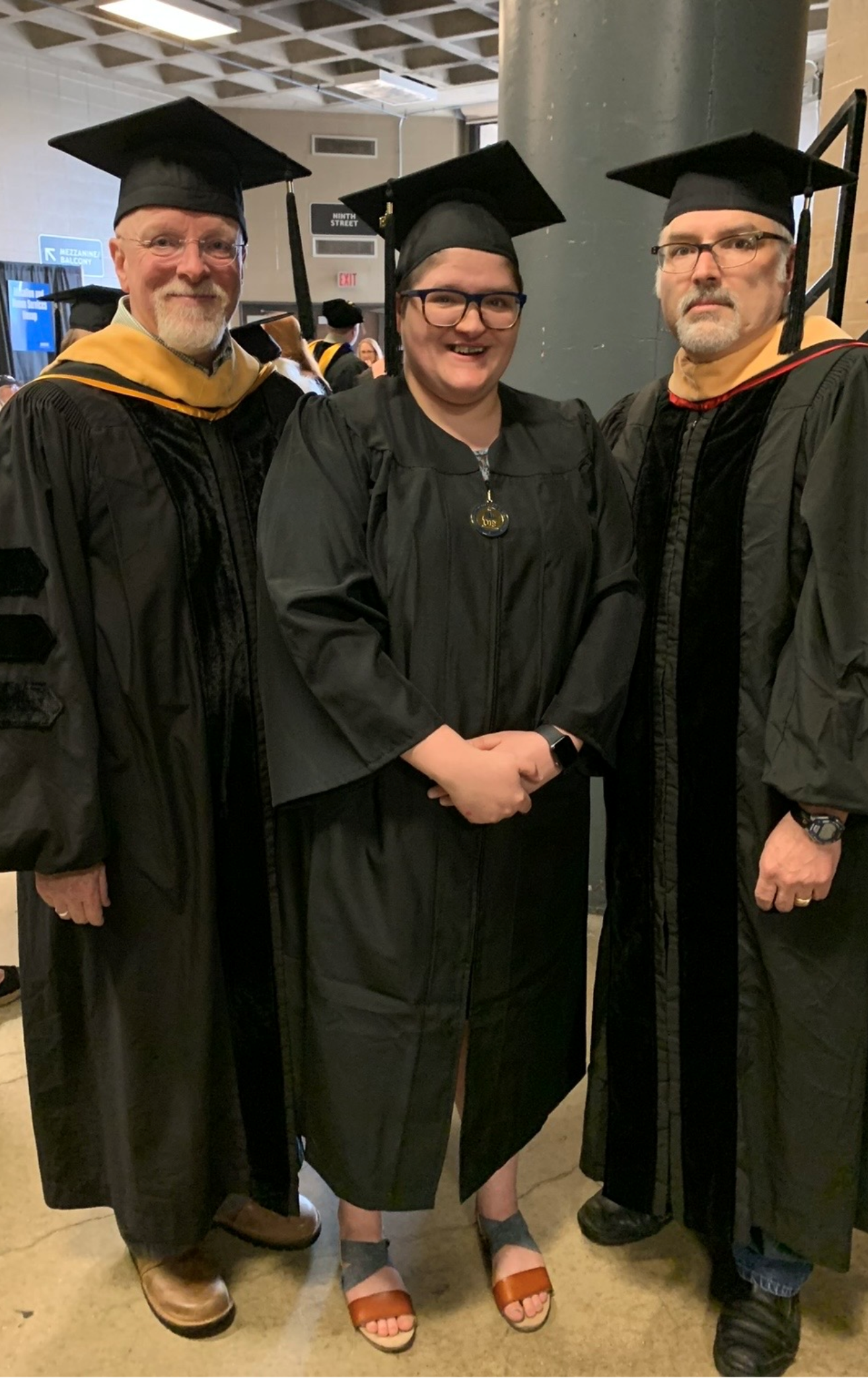 Three people in graduation gowns and caps, smiling indoors.