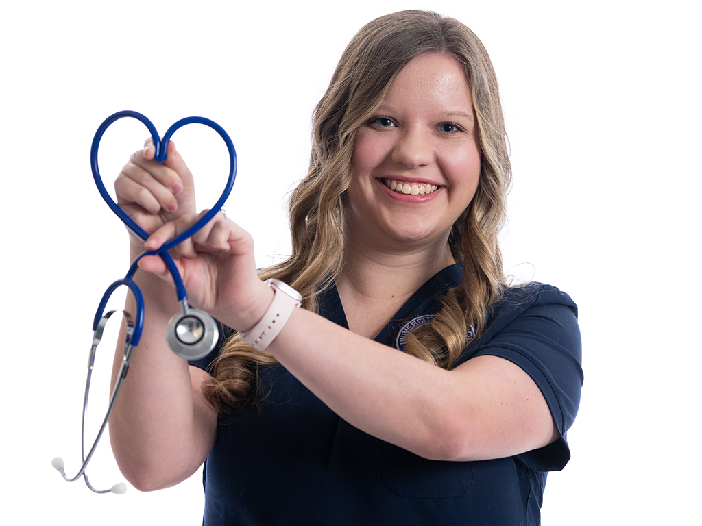 A smiling woman with long hair holds a stethoscope shaped into a heart, wearing a navy-blue shirt. The image conveys a sense of warmth and care.