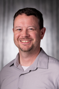 Smiling man in a gray shirt with a dark background.
