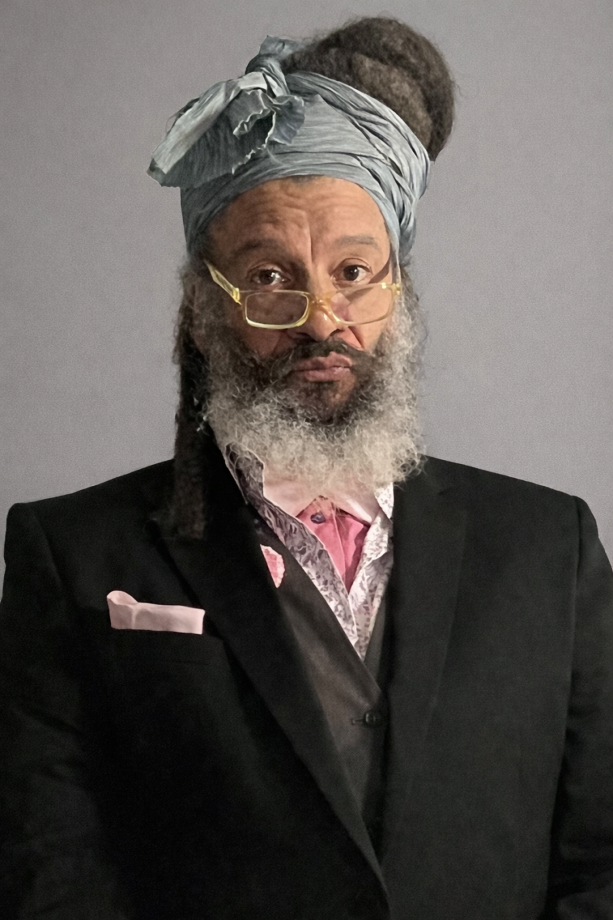 Man with beard and turban, wearing glasses and a black suit with a pink tie.
