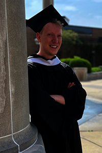 Graduate in cap and gown standing by a column.