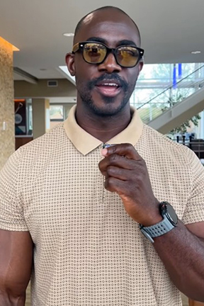 Man in a beige polo and sunglasses indoors, gesturing with hand.