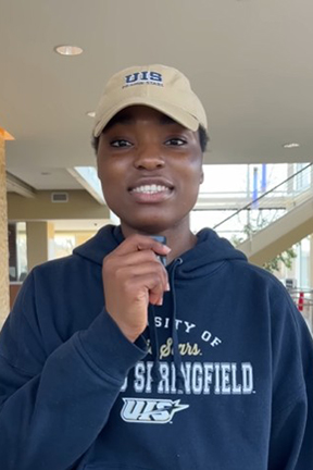 Young person indoors wearing a beige cap and navy hoodie, smiling.