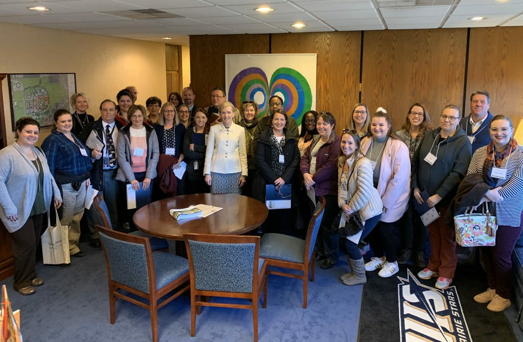 Employees are standing around a conference table posing for a picture, with the Chancellor in the middle.