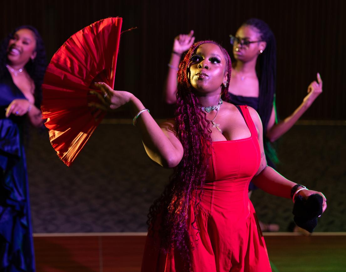 A woman in a red dress, waving a red fan, on a dark dance floor.