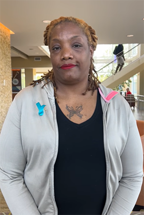 Woman with a neutral expression, wearing a gray hoodie with a blue and pink ribbon, indoors.