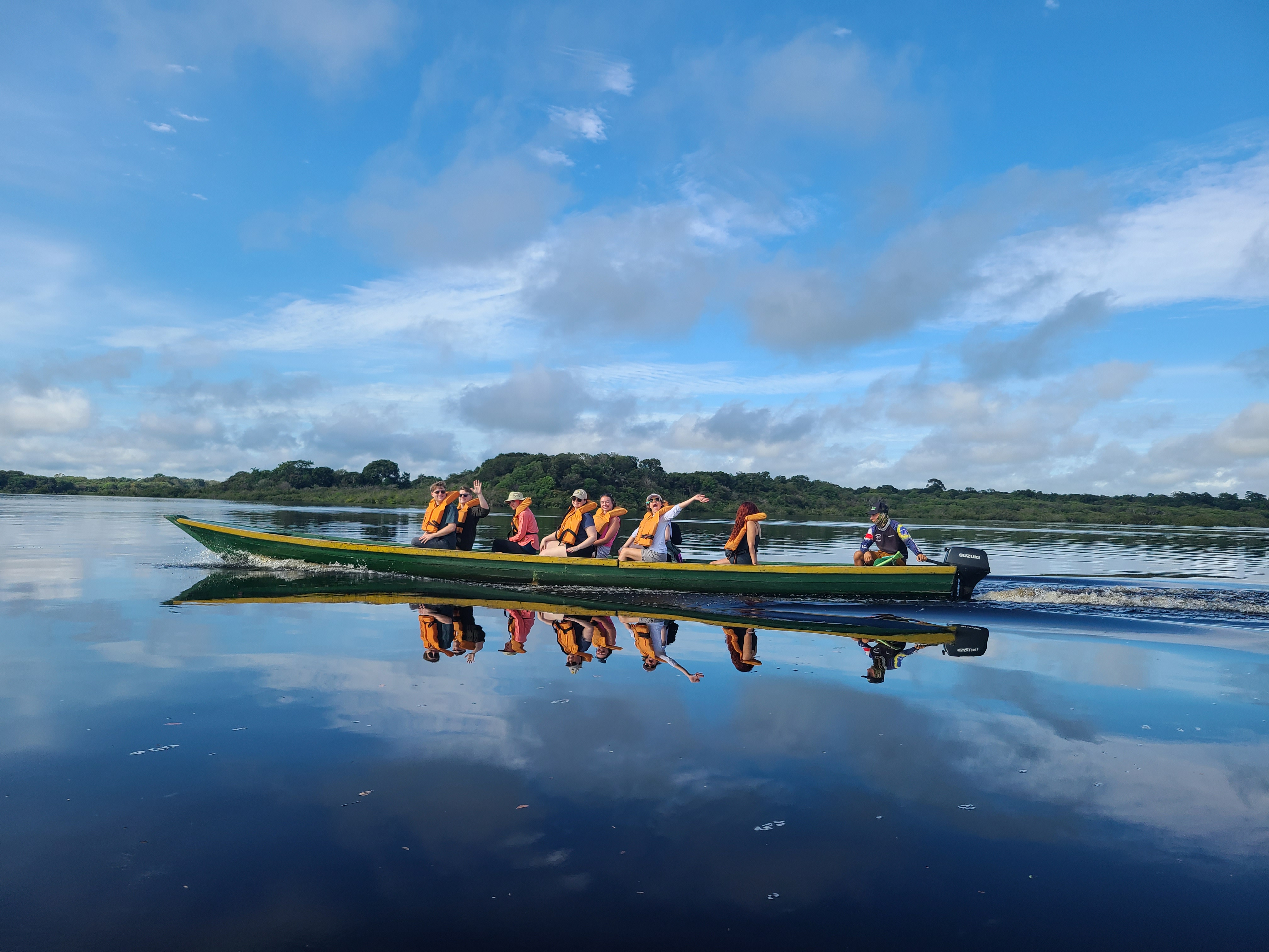 UIS students ride a boat across a calm Amazon River, with reflections mirrored on the water.