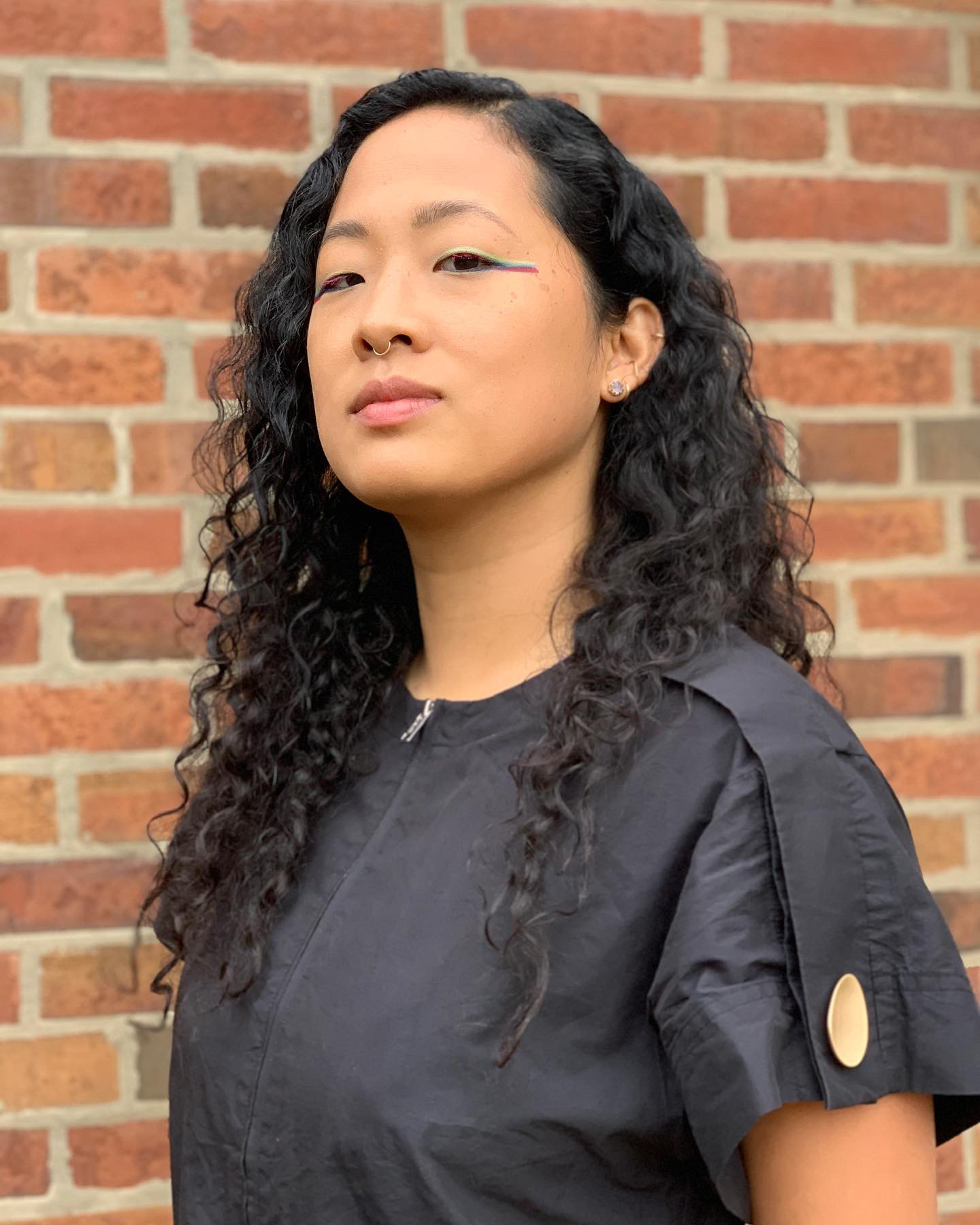 Portrait of artist Aram Han Sifuentes with long curly hair, colorful eyeliner, and a black top, posed against a red brick wall.