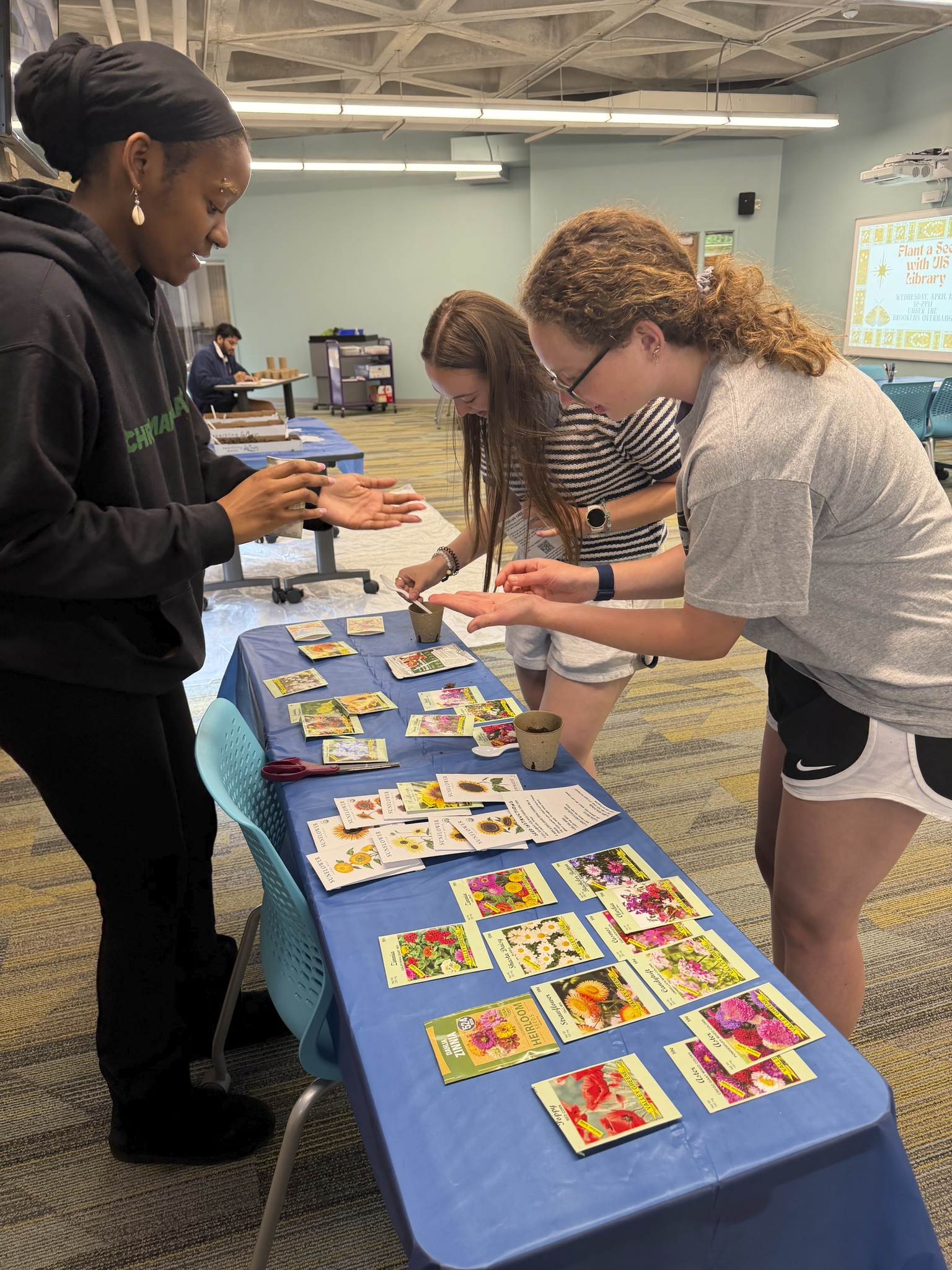 Library staff and students standing around table covered with seed packets and planting materials at Library "Plant a Seed" event.