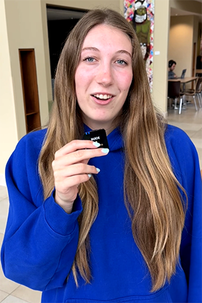 Young woman in a blue hoodie holding a small black object indoors.