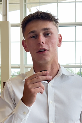 Young man in a white shirt speaking, large windows behind.