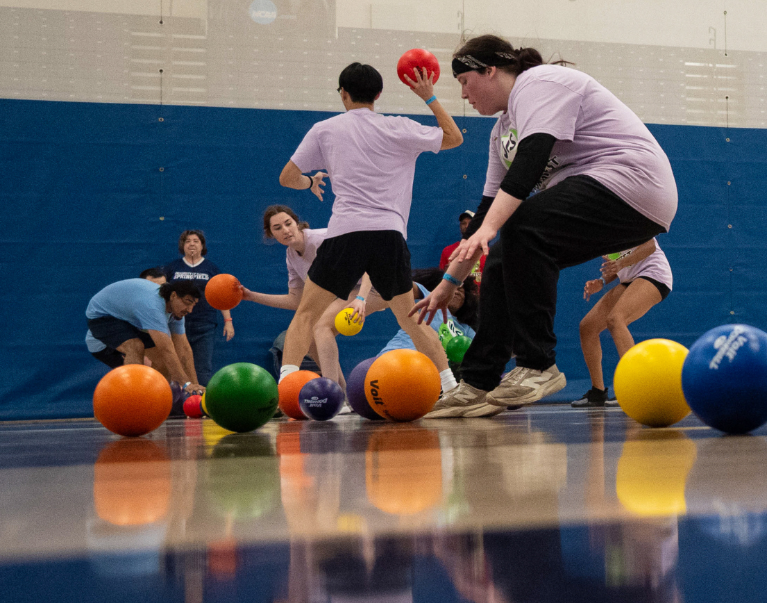 People in purple shirts running for colorful balls on a gym floor.