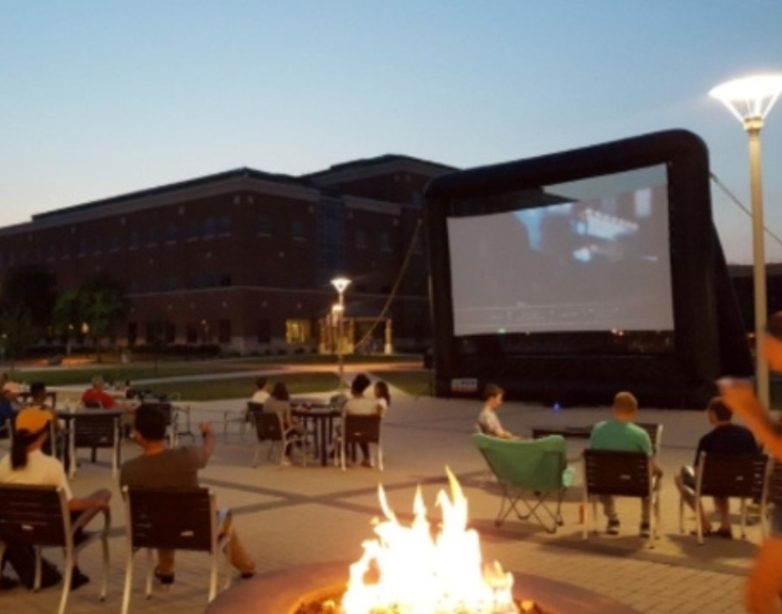 People gathered in lawn chairs on a patio to watch a projected screen. There is a large bonfire in the foreground.