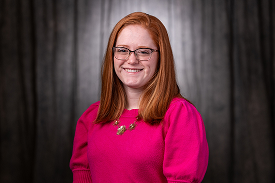 Smiling person with red hair and glasses, wearing a pink sweater, against a dark backdrop.