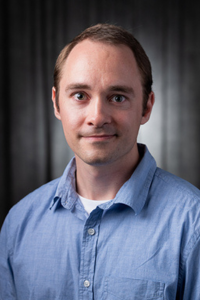 Man in a blue shirt against a dark background, smiling slightly.