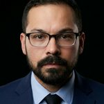 Man with glasses and beard wearing a suit in dark lighting.