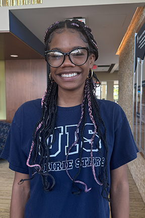 Smiling person with glasses and braided hair, wearing a navy blue shirt indoors.