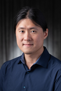 Man with dark hair, wearing a navy shirt, against a dark background.