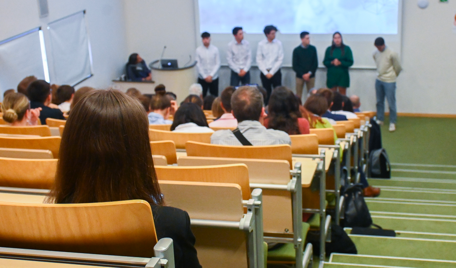 A view from the back of a lecture hall at IHECS in Belgium. Six students standing at the front while others are sitting in wooden lecture chairs.