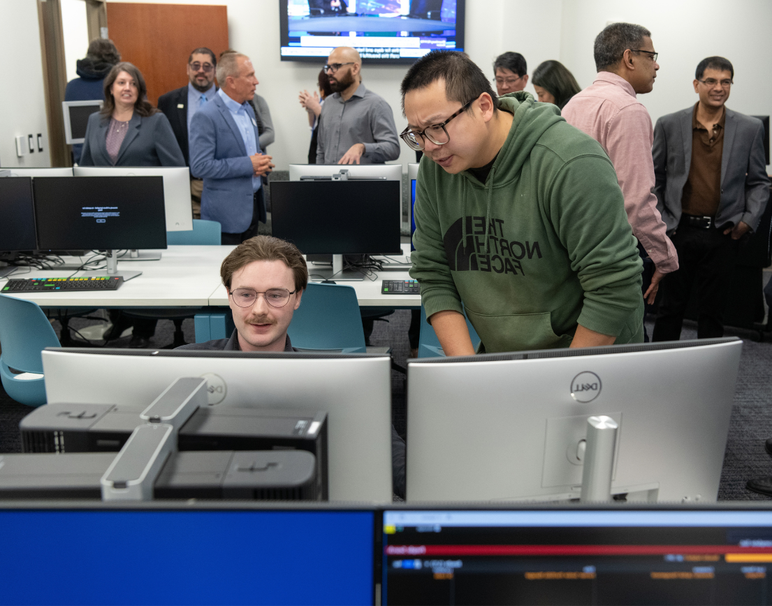 Two students looking over a computer with others in the background in a computer lab.