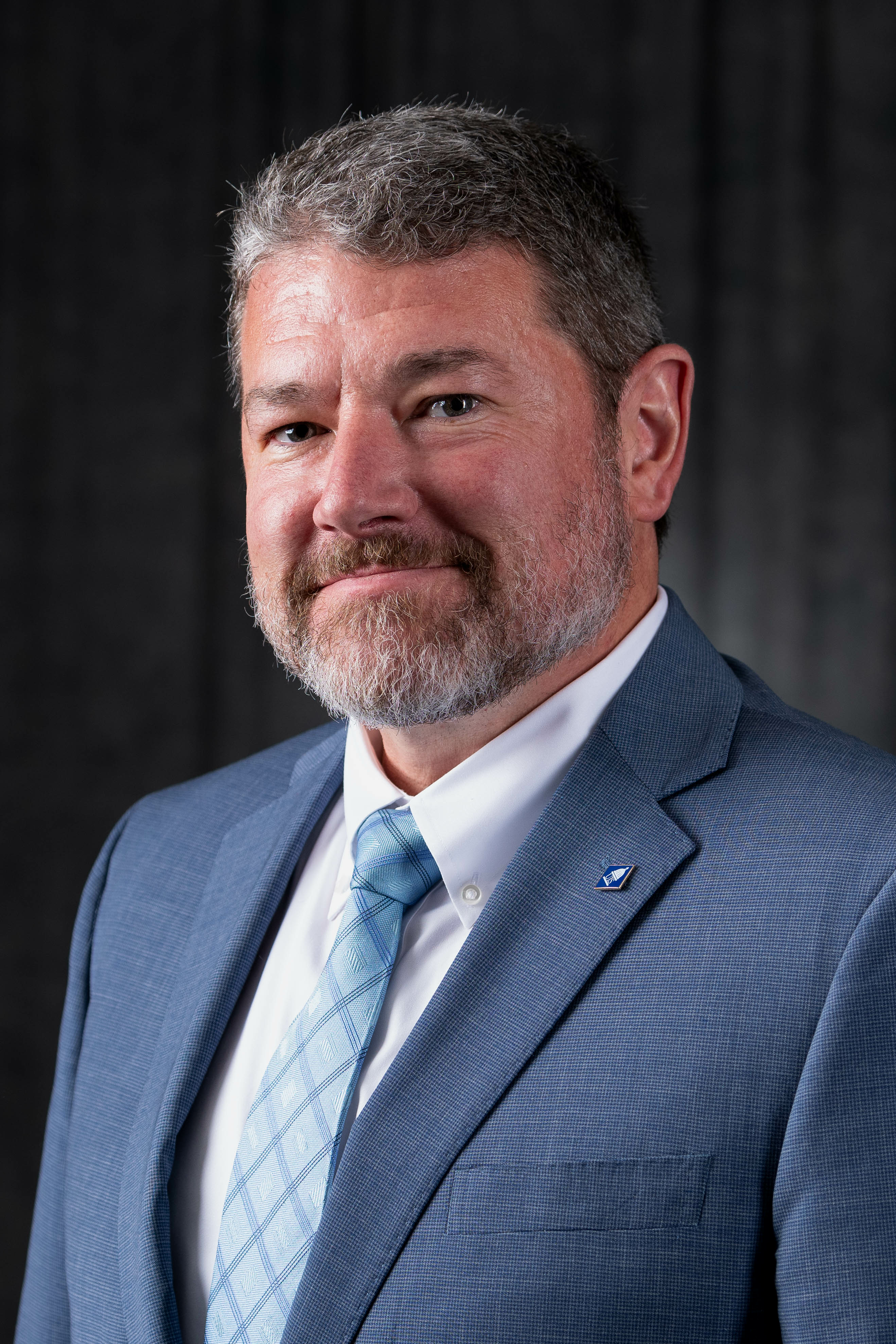 Head and shoulders photo of UIS Provost Brandon Schwab, wearing a medium jacket, white shirt, and light blue tie against a dark gray background.